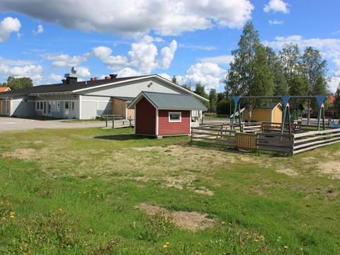 Sommarbild av grå träbyggnad, röd förrådsbod och en lekplats, med äng i förgrunden.