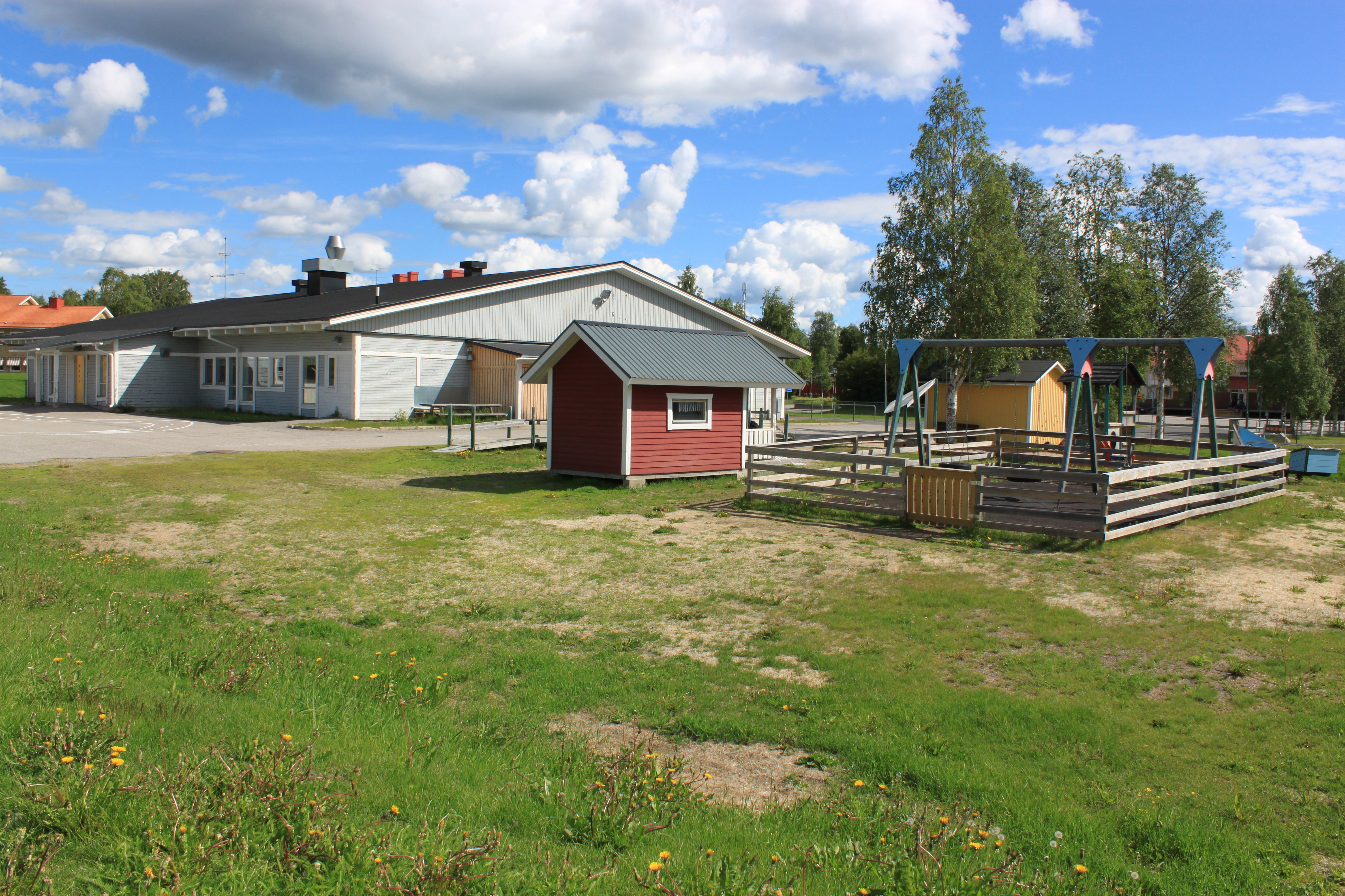 Sommarbild av grå träbyggnad, röd förrådsbod och en lekplats, med äng i förgrunden.