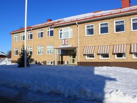 Vinterbild av tegelbyggnad med brunrandiga markiser och en flaggstång framför ingången