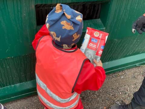 Bild som visar barn i röda kläder som sopsorterar mjölkpaket i grön container