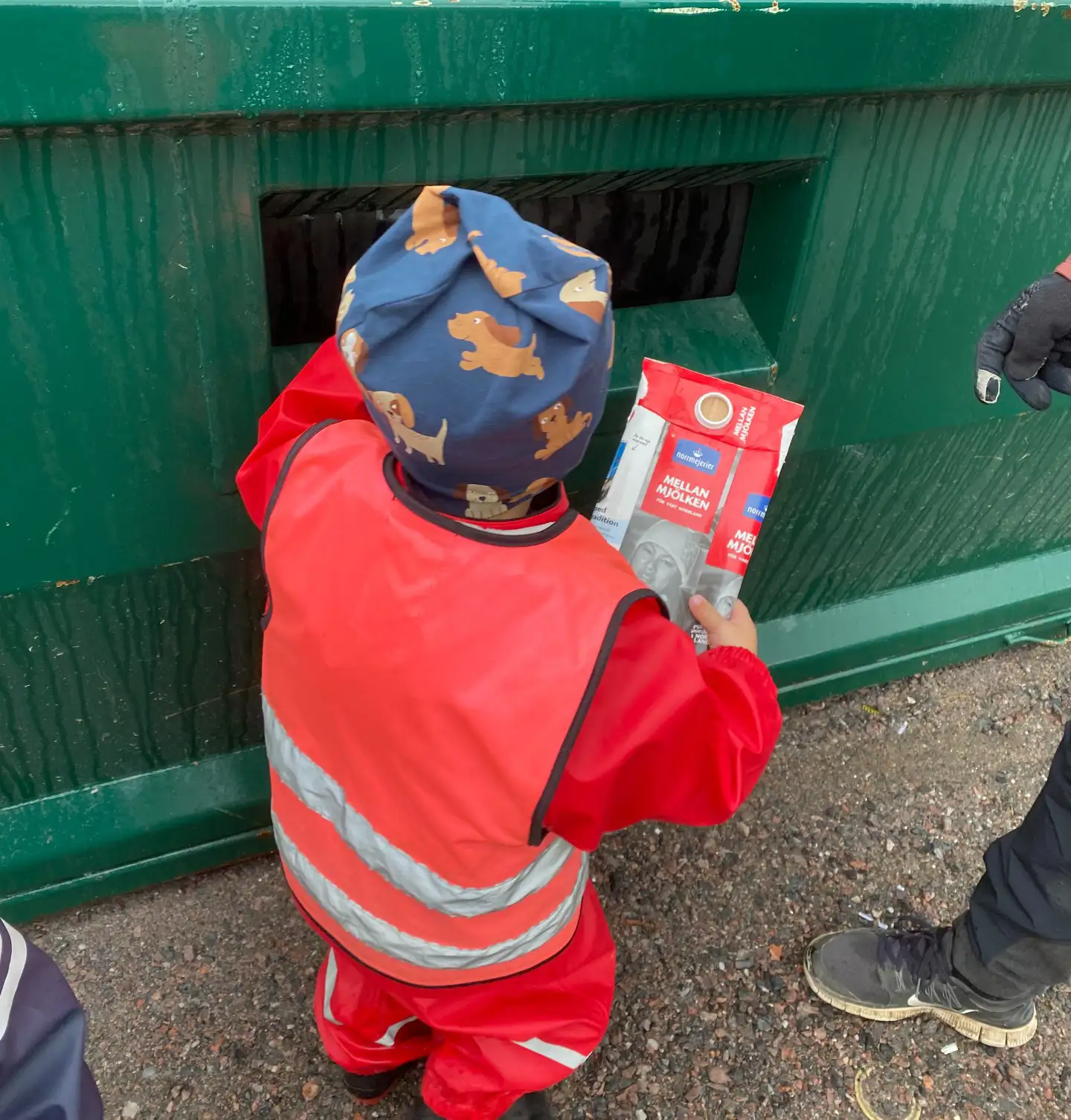 Bild som visar barn i röda kläder som sopsorterar mjölkpaket i grön container
