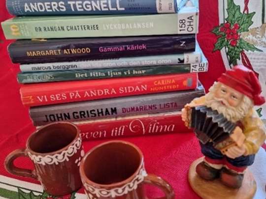 En trave med böcker, en keramiktomte och två glöggmuggar på en julduk.