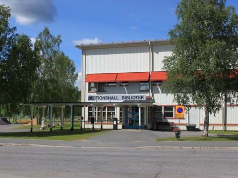 Sommarbild av ingång till vit träbyggnad i två våningar med röda markiser, björkar på vardera sidan av ingången.