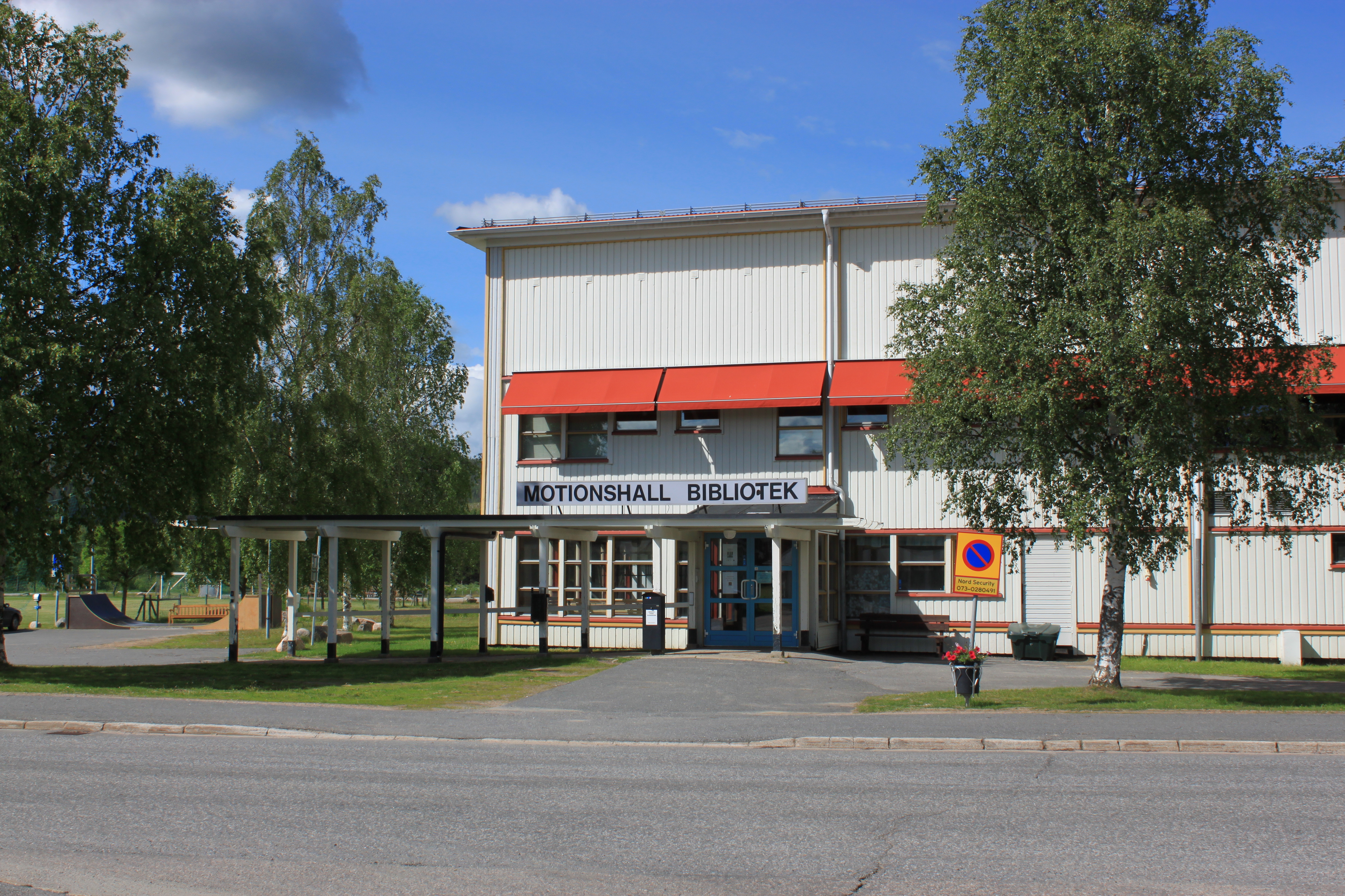 Sommarbild av ingång till vit träbyggnad i två våningar med röda markiser, björkar på vardera sidan av ingången.