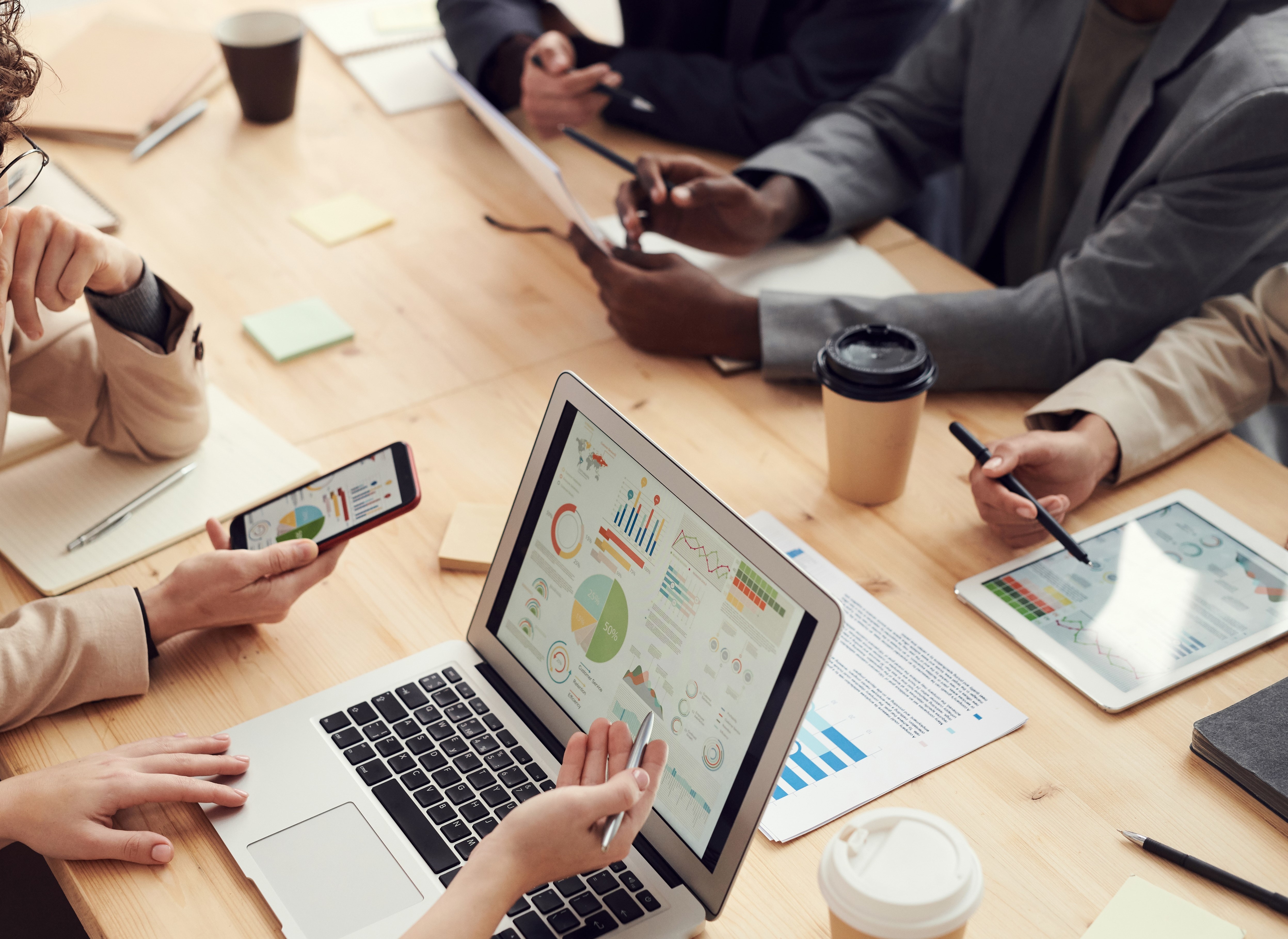 Flera människor som sitter runt ett skrivbord med datorer, plattor, mobiler och anteckningar.