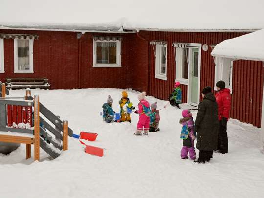 Bild som visar förskolegård med snö och lekande barn och personal omgivna av den röda förskolebyggnaden