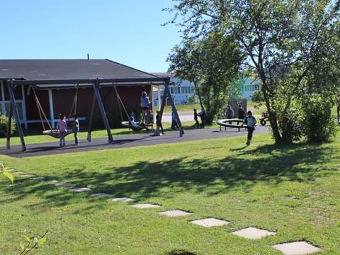 Sommarbild av lekpark med lekande barn, röd träbyggnad i bakgrunden.