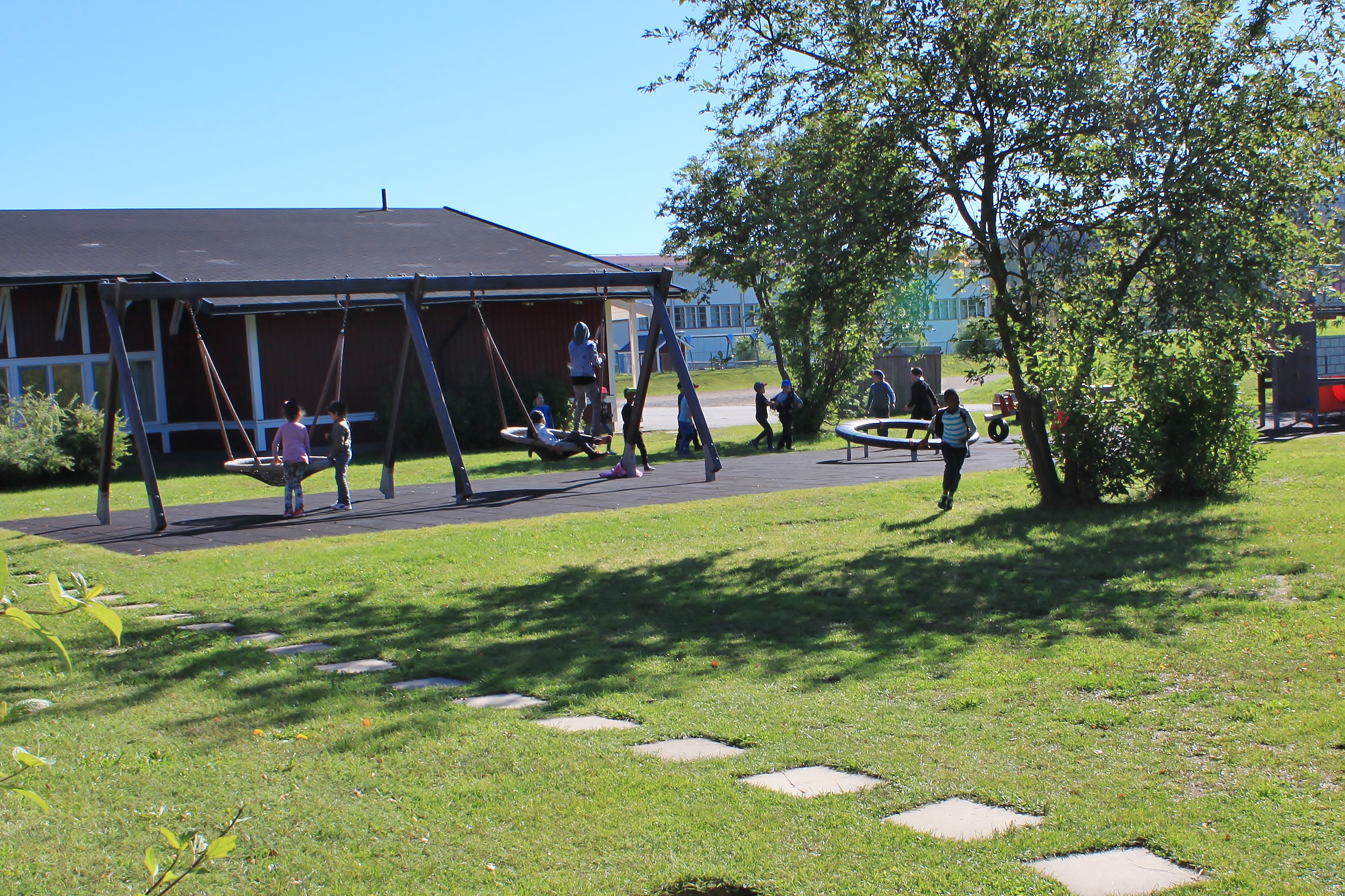 Sommarbild av lekpark med lekande barn, röd träbyggnad i bakgrunden.