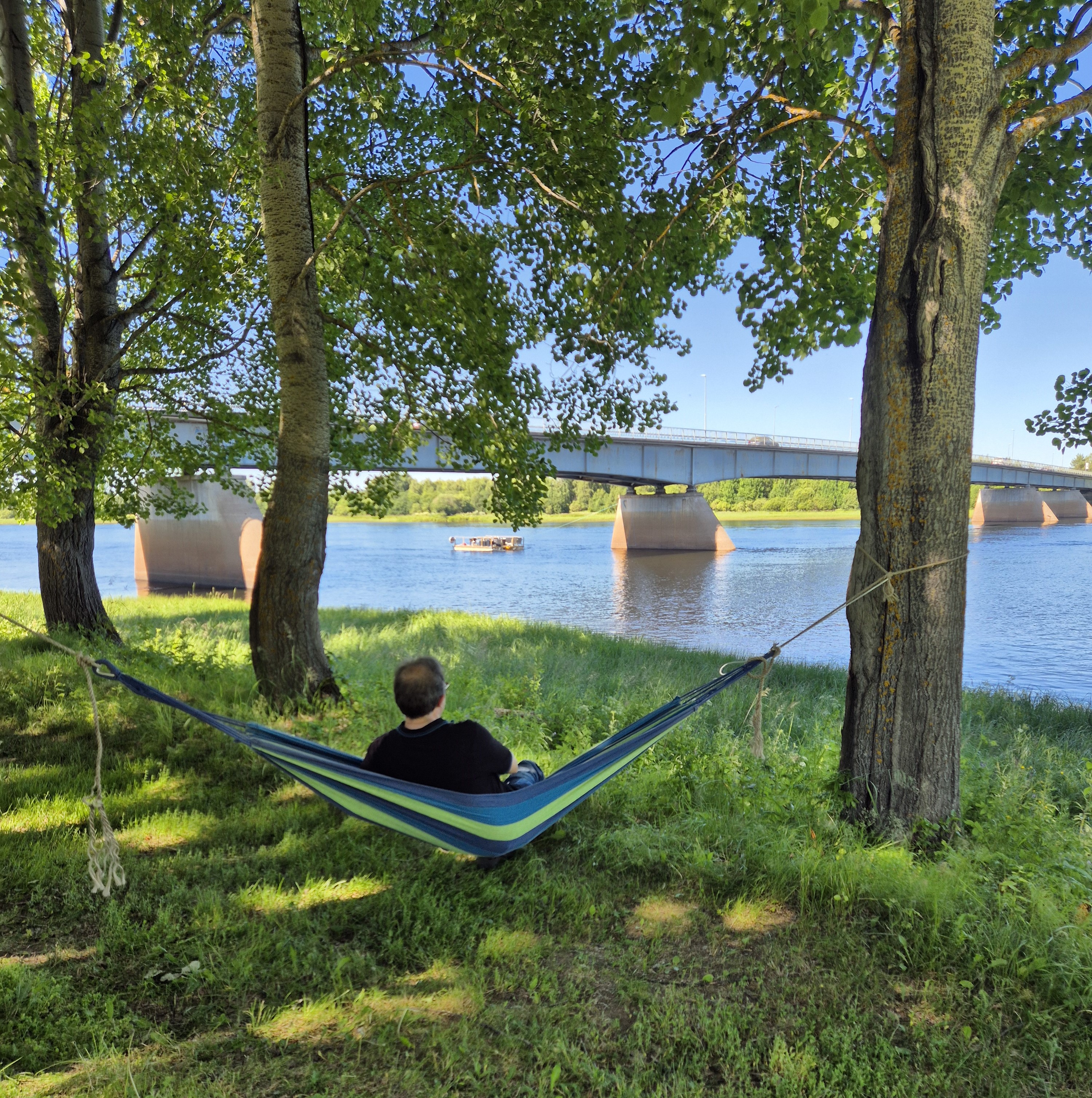 man sitter i en hängmatta mellan två grönskande träd och Kalix älv framför sig