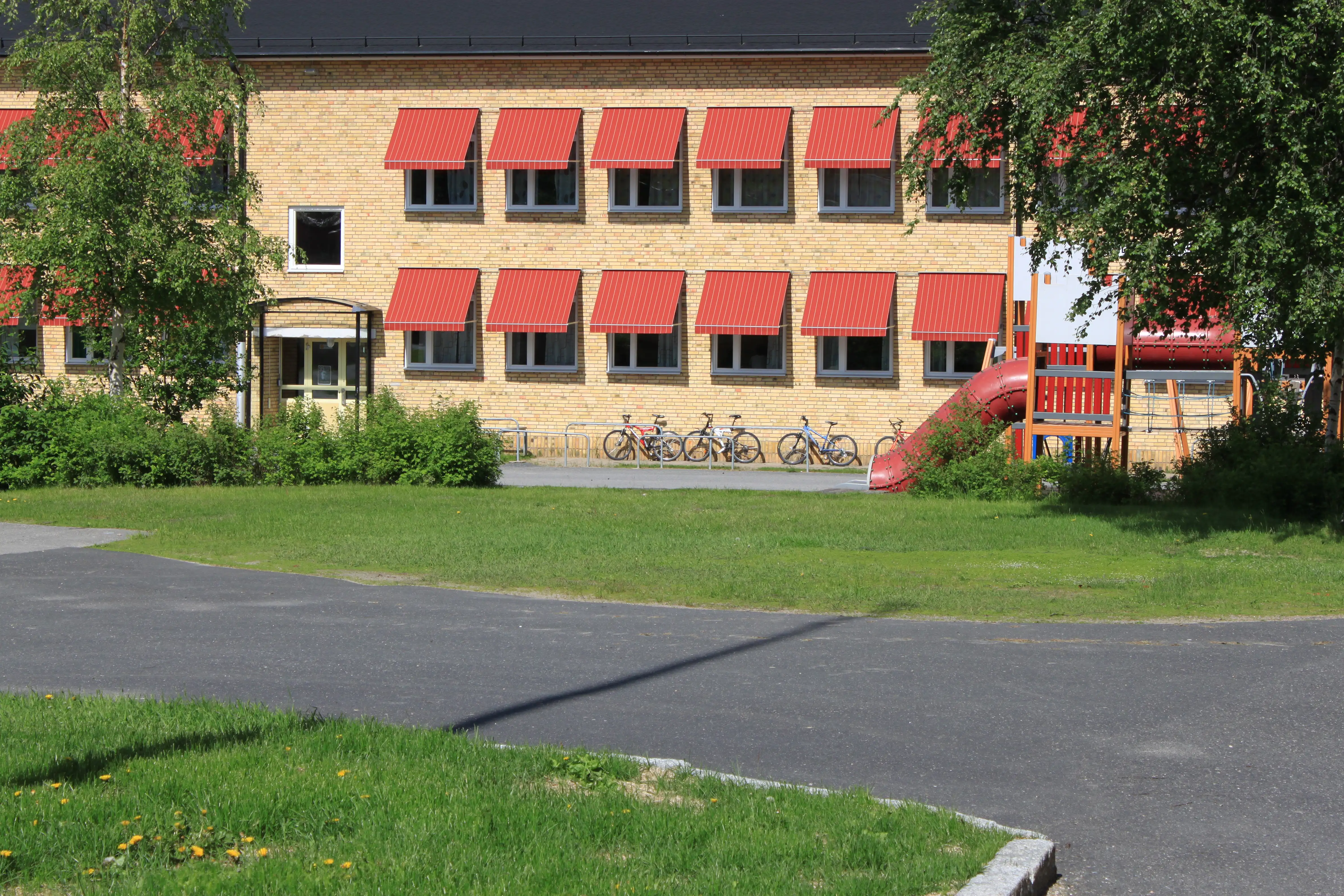 Sommarbild skolgård och tegelbyggnad med röda markiser i bakgrunden.